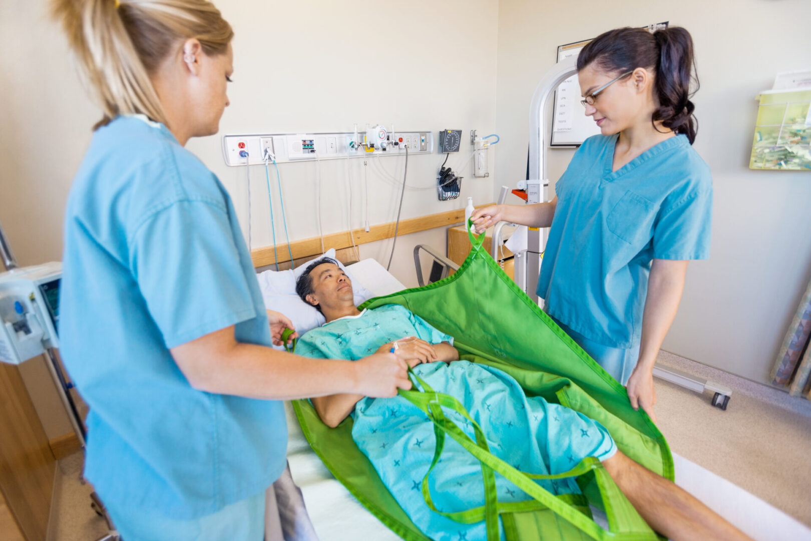 Caucasian nurses preparing male patient before transferring him on hydraulic lift at hospital room